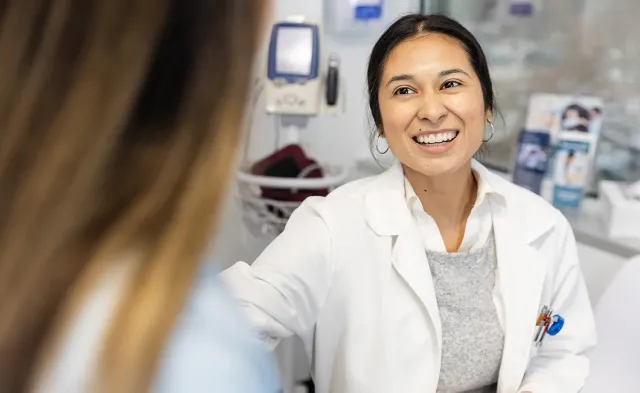 Smiling nurse practitioner in a white coat speaking with a patient in an exam room while holding a clipboard.