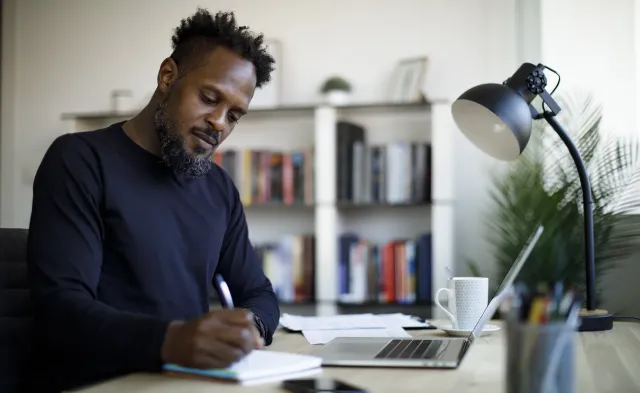 A student writes in a notebook at a desk with a laptop, phone, lamp, and bookshelves nearby.