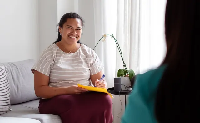 A smiling social worker and former RN sits on a couch holding a notepad and pen during a conversation with another person in a bright, comfortable room.