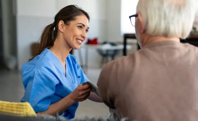 Smiling home health nurse in blue scrubs checks an older adult’s blood pressure during a patient care visit.