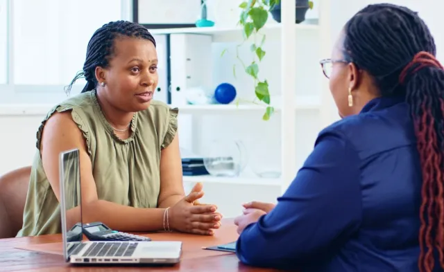 A human services worker speaking with a client in an office, engaged in a supportive one-on-one conversation with a laptop and office shelves in the background.