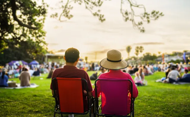 Couple sitting on the picnic chairs, listening to the music concert. Unrecognizable crowd of people in the park.