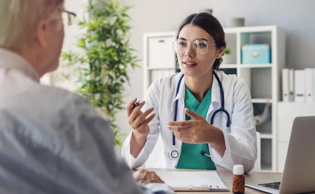 Nurse with a DNP wearing a stethoscope discusses care with an older patient during an office visit, seated across a desk with a laptop and paperwork.