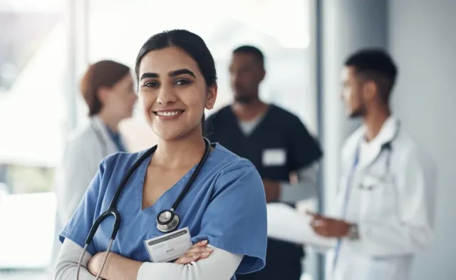 RN in blue scrubs with a stethoscope smiles confidently at the camera while colleagues collaborate in the background of a clinical setting.