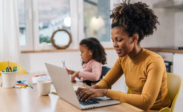 Parent works on a laptop at a kitchen table while a young child sits nearby doing arts and crafts, balancing work and family responsibilities at home.