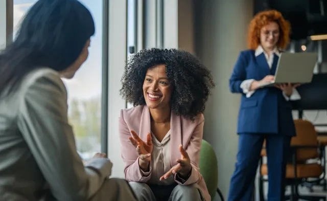 Three behavioral health professionals converse in a modern office, with one woman smiling and gesturing while another listens and a third works on a laptop in the background.