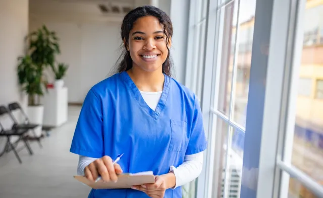 A smiling nurse in blue scrubs holds a clipboard and pen while standing beside large windows in a bright healthcare facility.