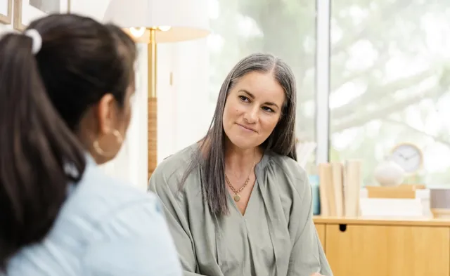A holistic nurse listens attentively to a patient while holding a notepad and pen during a one-on-one conversation in a calm setting.
