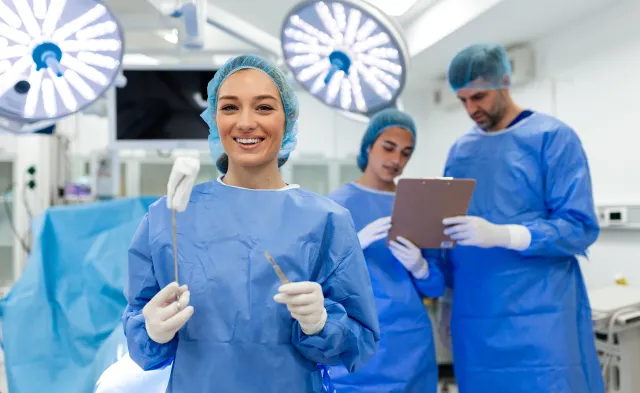 ortrait of Smiling Female Surgeon in Blue Scrubs Holding Surgical Scalpel and Medical Forceps in Modern Operating Room with Professional Medical Team in Background