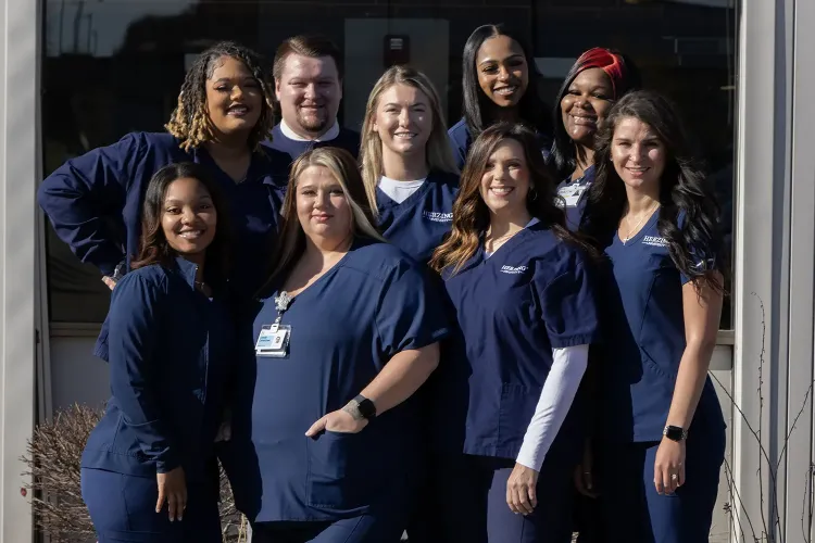 Herzing University practical nursing graduates wearing blue scrubs smiling outside of campus.