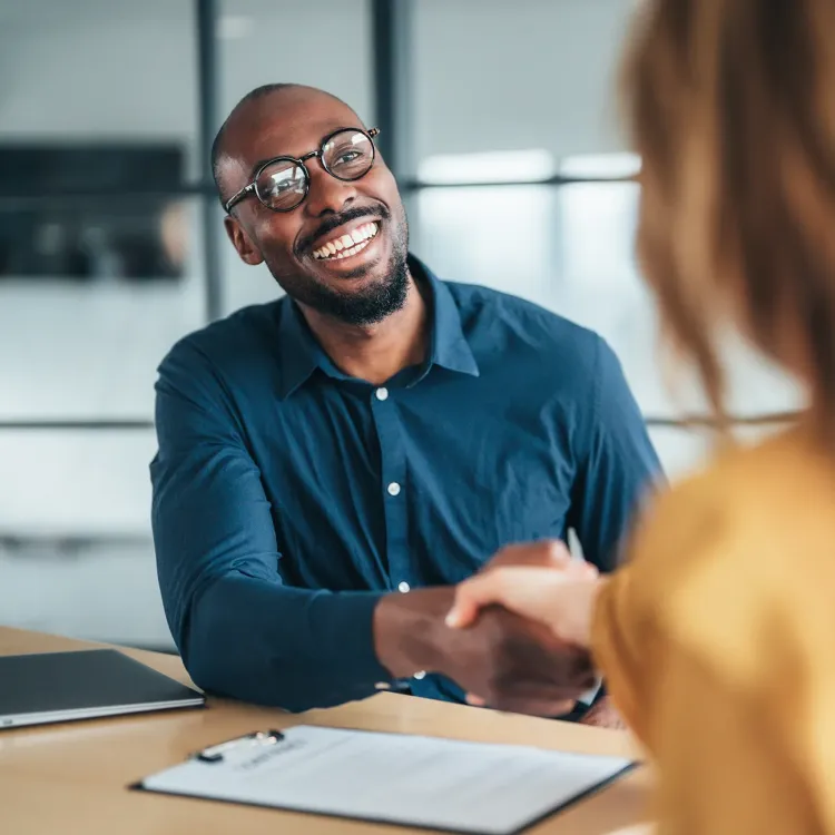 A smiling financial consultant shakes hands with another person across a desk during a meeting.