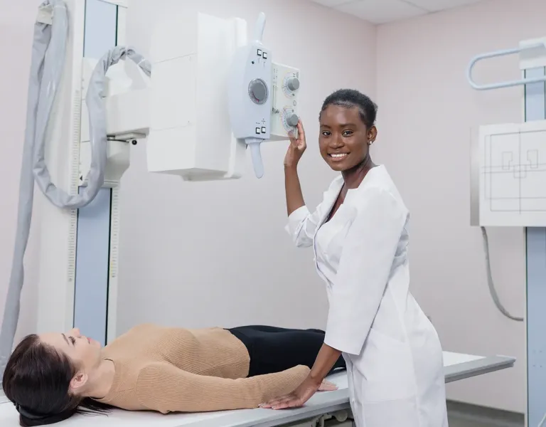 A radiologic technologist in a white lab coat adjusts an X-ray machine while standing beside a patient lying on the examination table. The technologist is smiling and looking at the camera, and the patient appears relaxed with her arms at her sides.