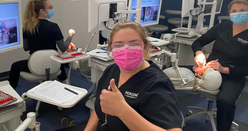 Herzing dental assisting student wearing mask and giving thumbs up while practicing teeth cleaning on dummy