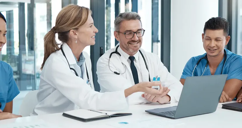 Group of healthcare professionals, including doctors and nurses, gathered around a table in a modern medical office, smiling and collaborating while reviewing information on a laptop