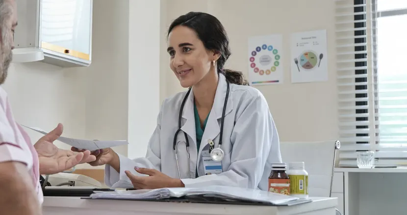 A female healthcare provider in a white coat and stethoscope smiles warmly while handing a paper to an older male patient during a consultation in a medical office