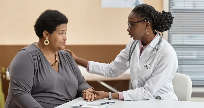 A nurse practitioner sits at a table with an adult patient, offering support by holding the patient's hand and placing a reassuring hand on her shoulder during a conversation in a medical office