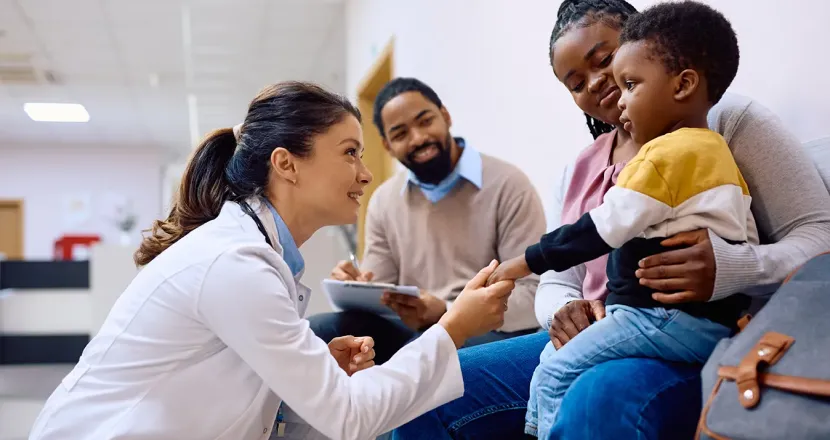 Smiling healthcare professional in a white coat kneeling and greeting a young boy sitting on his mother's lap in a medical waiting area, with the boy's father smiling in the background