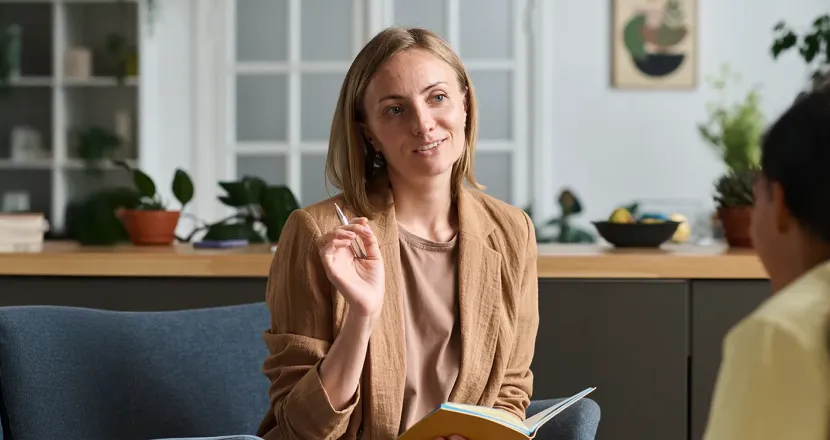 A social worker attentively listens and takes notes while engaging in a one-on-one counseling session with a client in a comfortable, plant-filled office setting