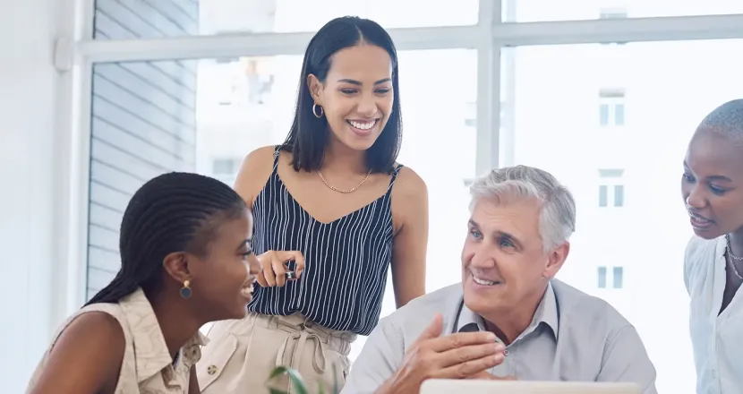 Group of four diverse colleagues gathered around a desk, smiling and engaged in a discussion, with one older man speaking while others listen attentively in a bright, modern office setting.
