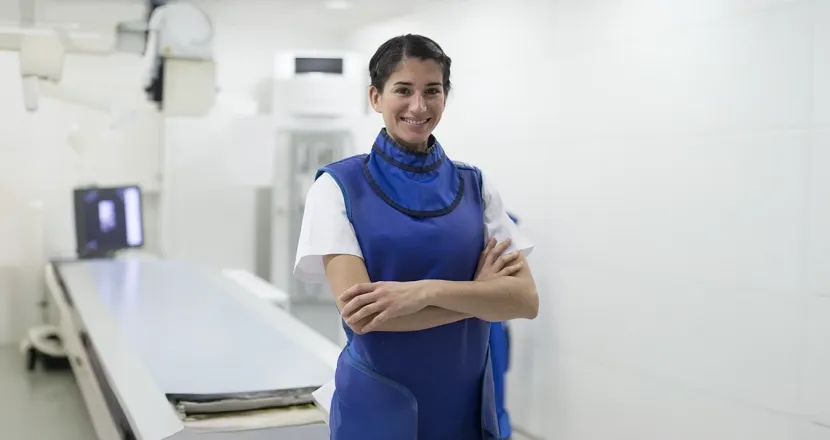 Radiologic technologist standing confidently in a clinical setting wearing a lead apron near medical imaging equipment.