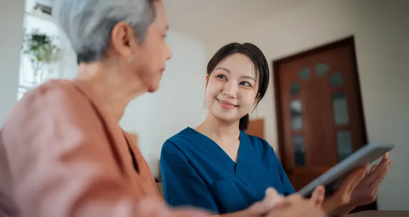 A healthcare worker in blue scrubs smiles warmly at an older woman seated beside her in a home setting, suggesting a supportive caregiving conversation.