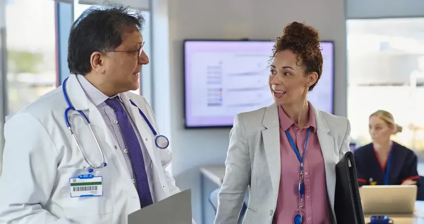 A healthcare administrator walks through a clinical training room with a doctor while nurses in scrubs work together at laptops in the background.