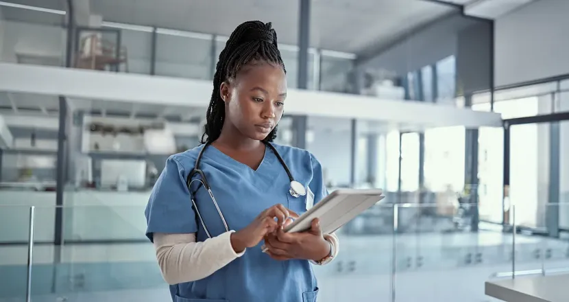 A nurse in blue scrubs uses a tablet in a bright clinical setting, reviewing digital information while standing near glass offices.