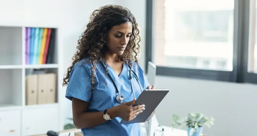 A healthcare professional in blue scrubs reviews information on a tablet while standing in a bright office with large windows.