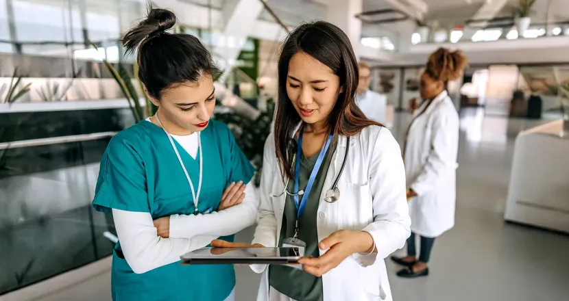 A nurse and clinician review information together on a tablet in a healthcare setting while other medical staff work in the background.