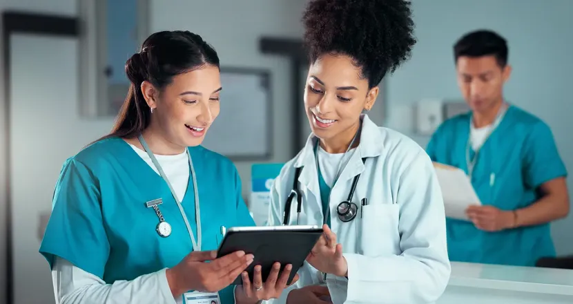 A nurse and clinician review information together on a tablet at a hospital workstation while another healthcare worker stands behind them.