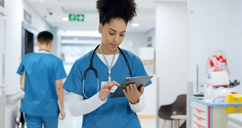 A nurse in blue scrubs reviews information on a tablet in a hospital hallway as another healthcare worker walks away in the background.