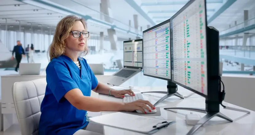 A nurse in blue scrubs works at a computer station with multiple monitors displaying healthcare data in a modern clinical setting.