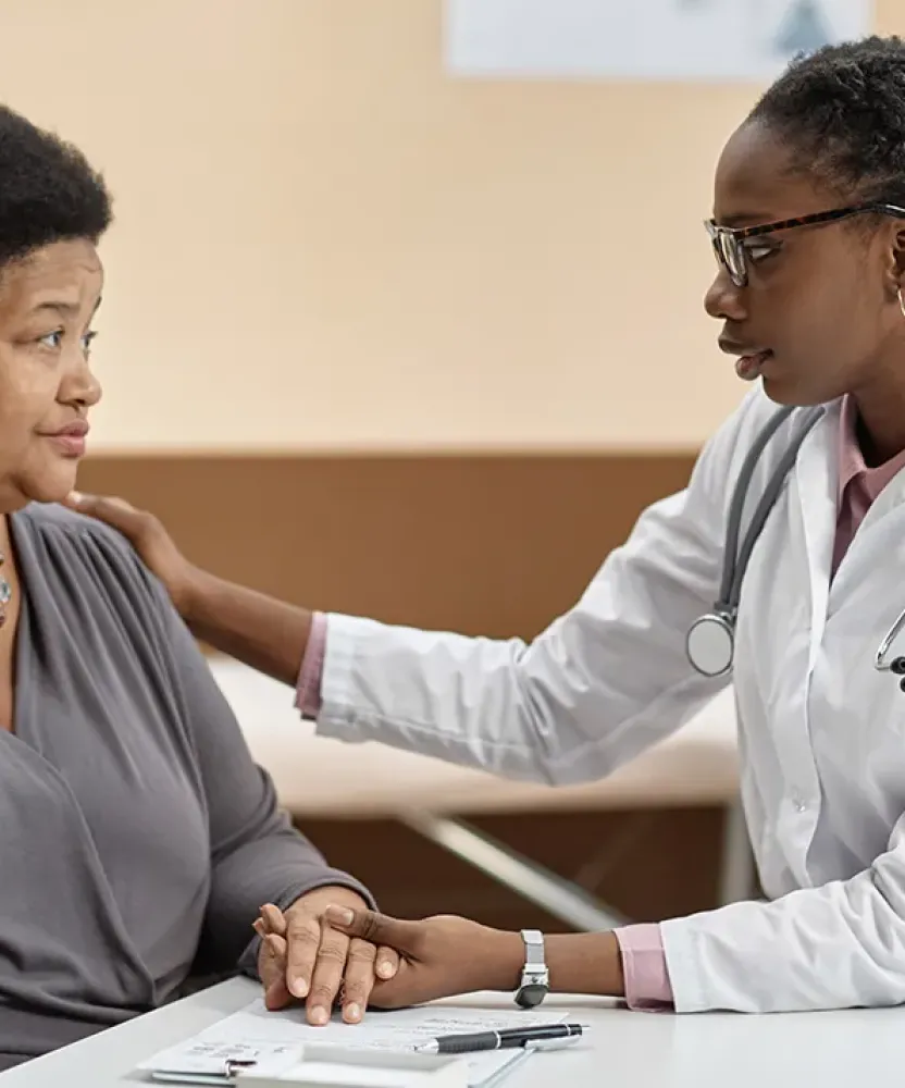 A psychiatric mental health nurse practitioner sits at a table with an adult patient, offering support by holding the patient's hand and placing a reassuring hand on her shoulder during a conversation in a medical office