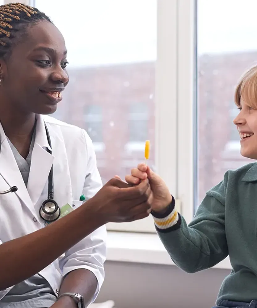 Woman pediatric primary care nurse practitioner smiling and handing a lollipop to a young boy in a clinic setting, with snow visible outside the window