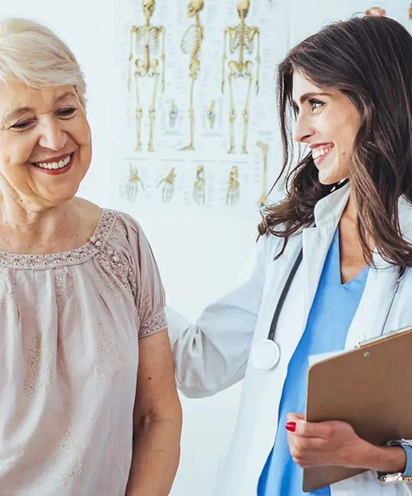Smiling women's health nurse practitioner wearing a white coat and stethoscope holding a clipboard, warmly interacting with an older female patient in a medical office