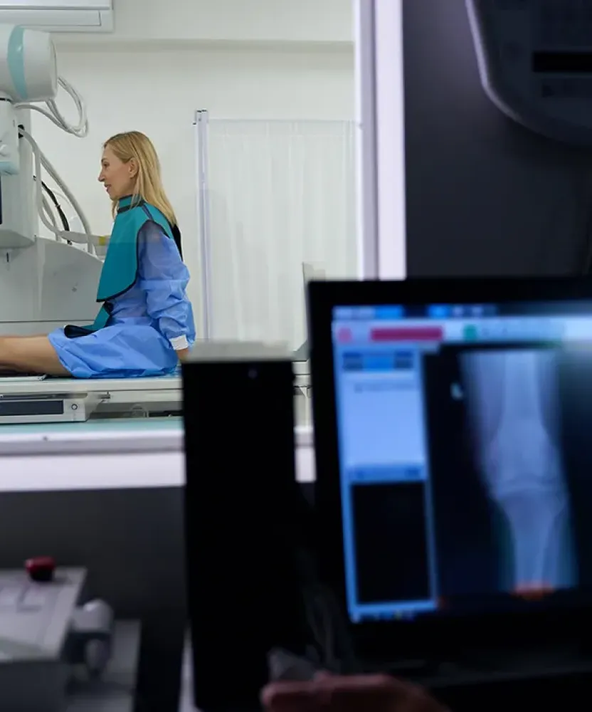 A Tampa radiologic technologist reviews X-ray images of a patient's leg on a computer screen in a dark control room, while the patient sits on a table in the adjacent imaging room.
