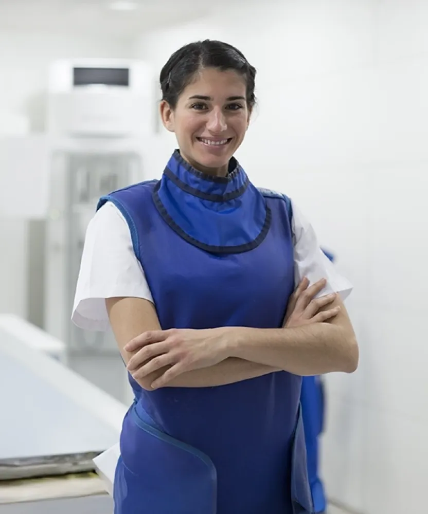 Radiologic technologist standing confidently in a clinical setting wearing a lead apron near medical imaging equipment.