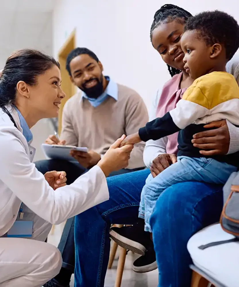Smiling family nurse practitioner kneeling and greeting a young boy sitting on his mother's lap in a medical waiting area, with the boy's father smiling in the background