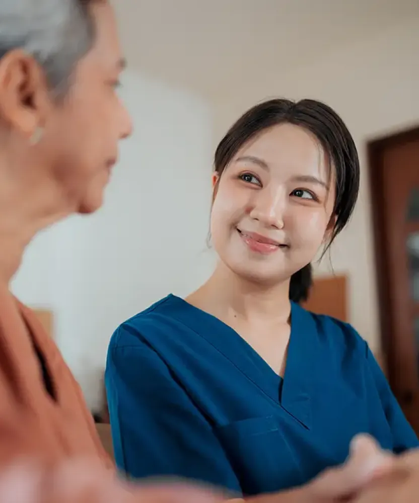 A holistic nurse in blue scrubs smiles warmly at an older woman seated beside her in a home setting, suggesting a supportive caregiving conversation.