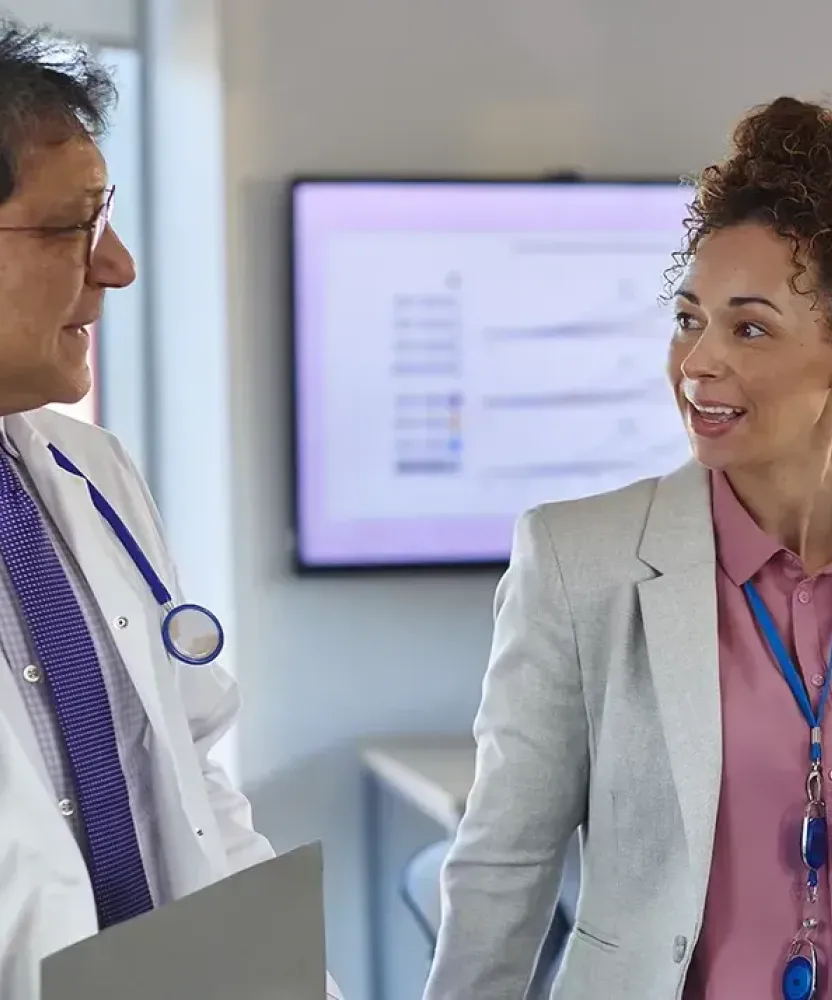 A nurse executive with an MSN and MBA walks through a clinical training room with a doctor while nurses in scrubs work together at laptops in the background.
