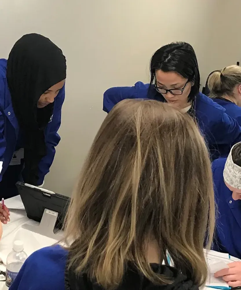 Herzing Minneapolis nursing students in blue scrubs gather around a medical training mannequin during a hands-on classroom skills activity.