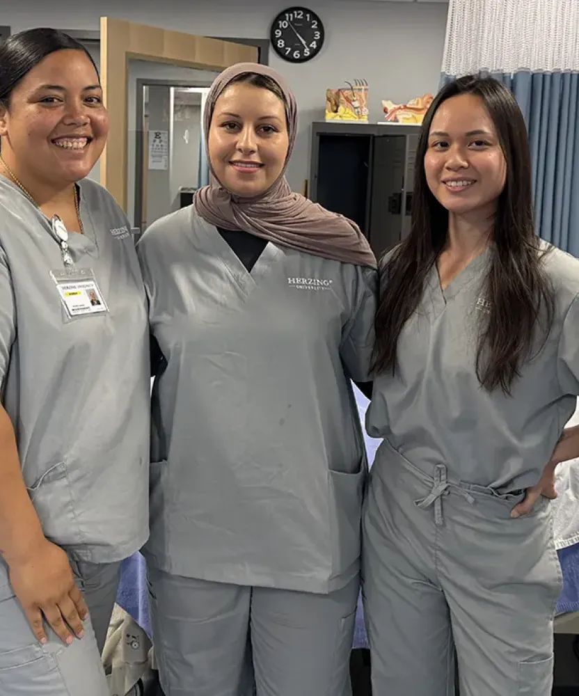 Three Herzing University BSN students wearing scrubs stand together in a simulated hospital room with a patient bed and medical equipment during nursing skills training.
