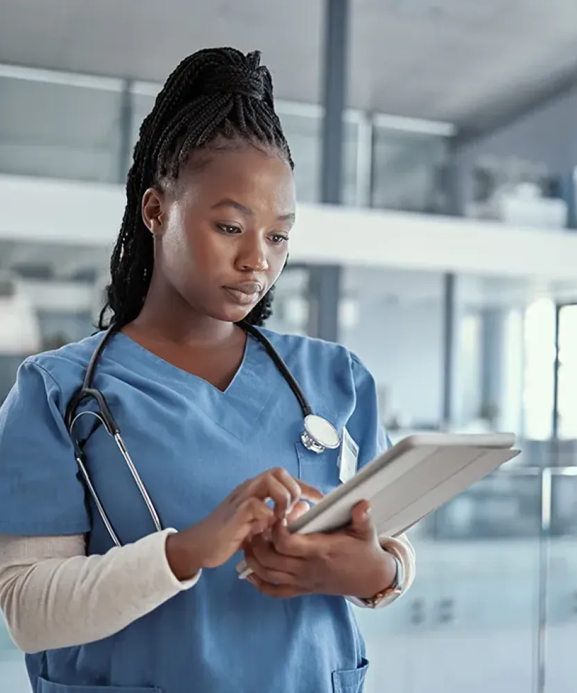 A nurse in blue scrubs uses a tablet in a bright clinical setting, reviewing digital information while standing near glass offices.