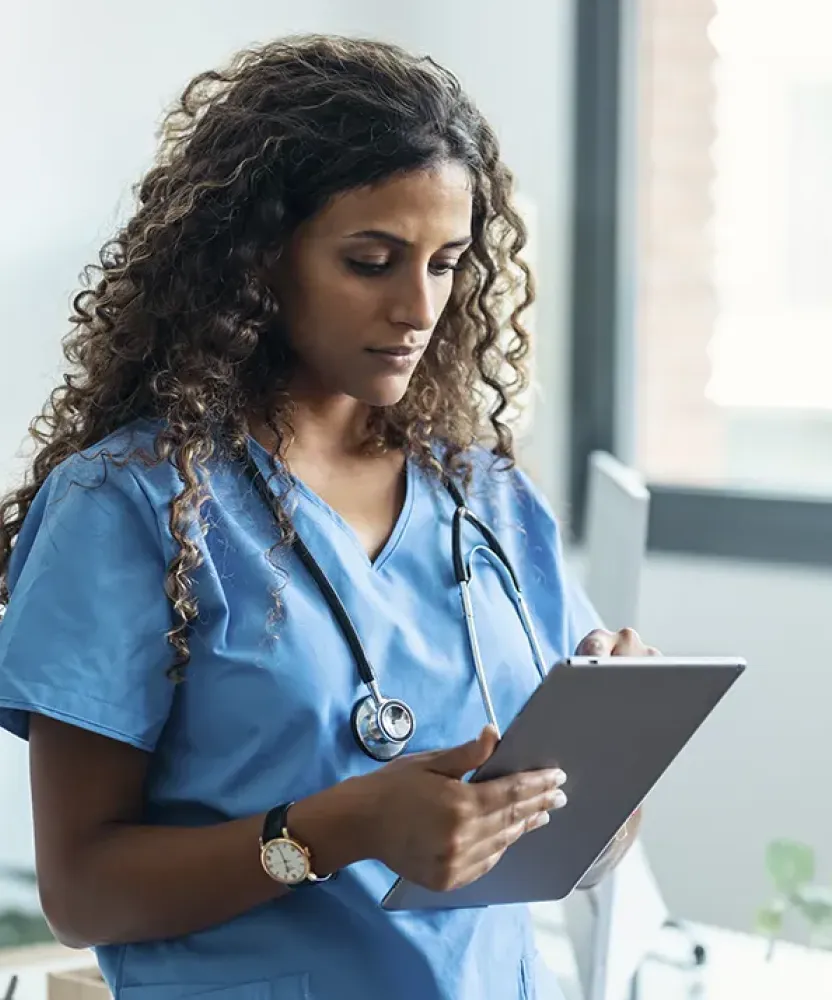 A healthcare professional in blue scrubs reviews information on a tablet while standing in a bright office with large windows.
