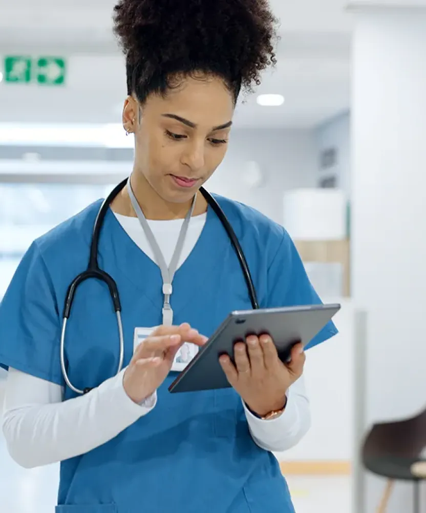 A nurse in blue scrubs reviews information on a tablet in a hospital hallway as another healthcare worker walks away in the background.