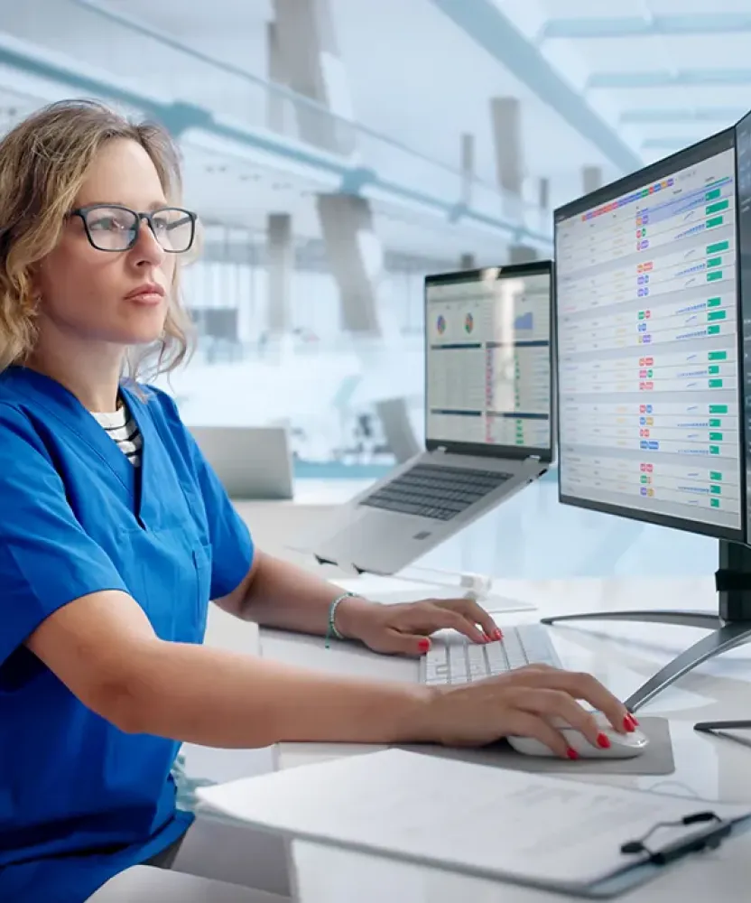 A nurse in blue scrubs works at a computer station with multiple monitors displaying healthcare data in a modern clinical setting.