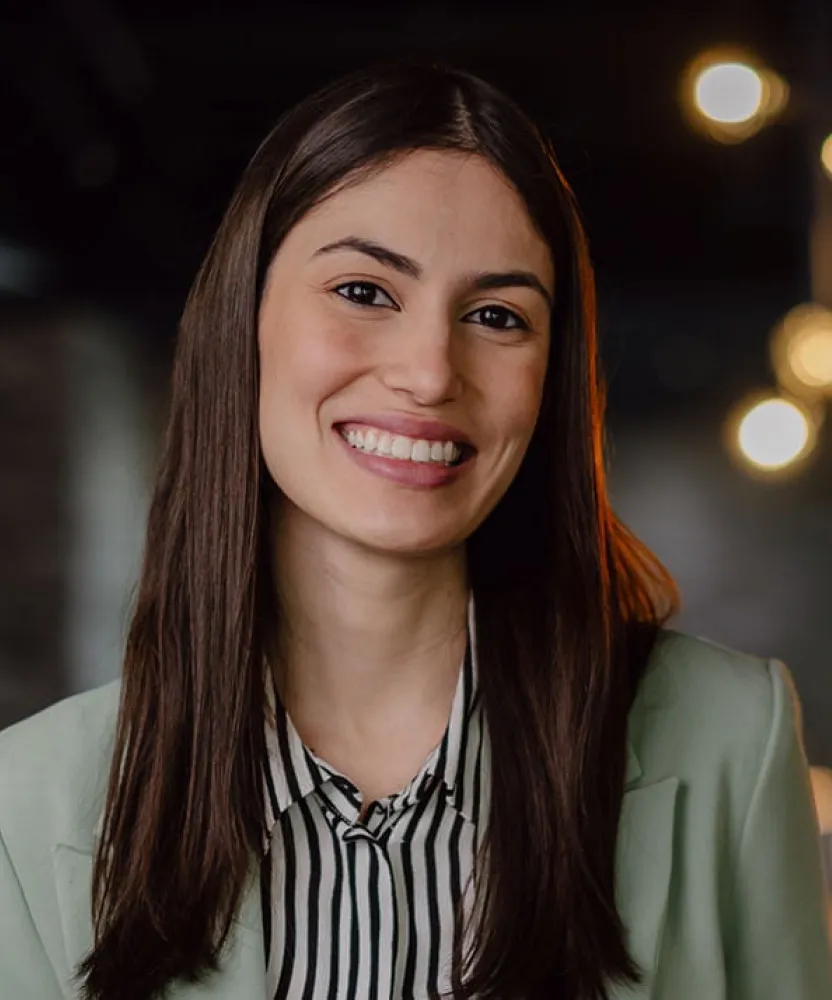 Smiling business professional standing in a modern office with coworkers gathered around a table in the background