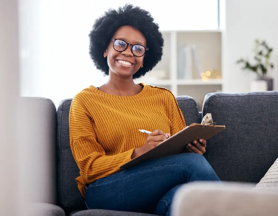 Smiling substance abuse counselor with natural hair and glasses sitting on a couch, holding a clipboard and pen during a therapy session.