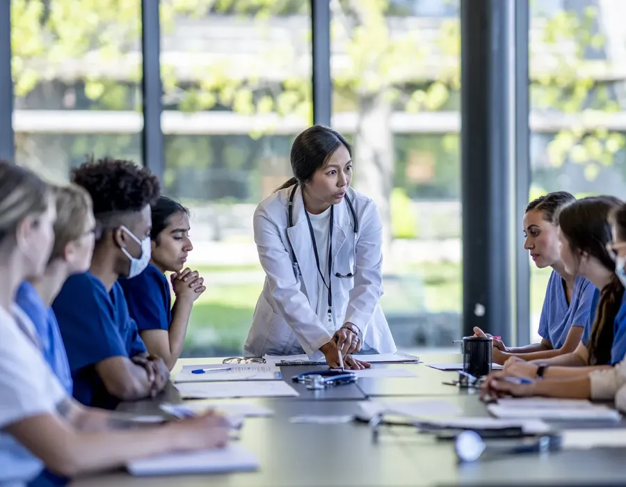 Nursing instructor in a white lab coat leads a group discussion with nursing students seated around a table in a bright classroom, reviewing medical charts and notes during a team learning session