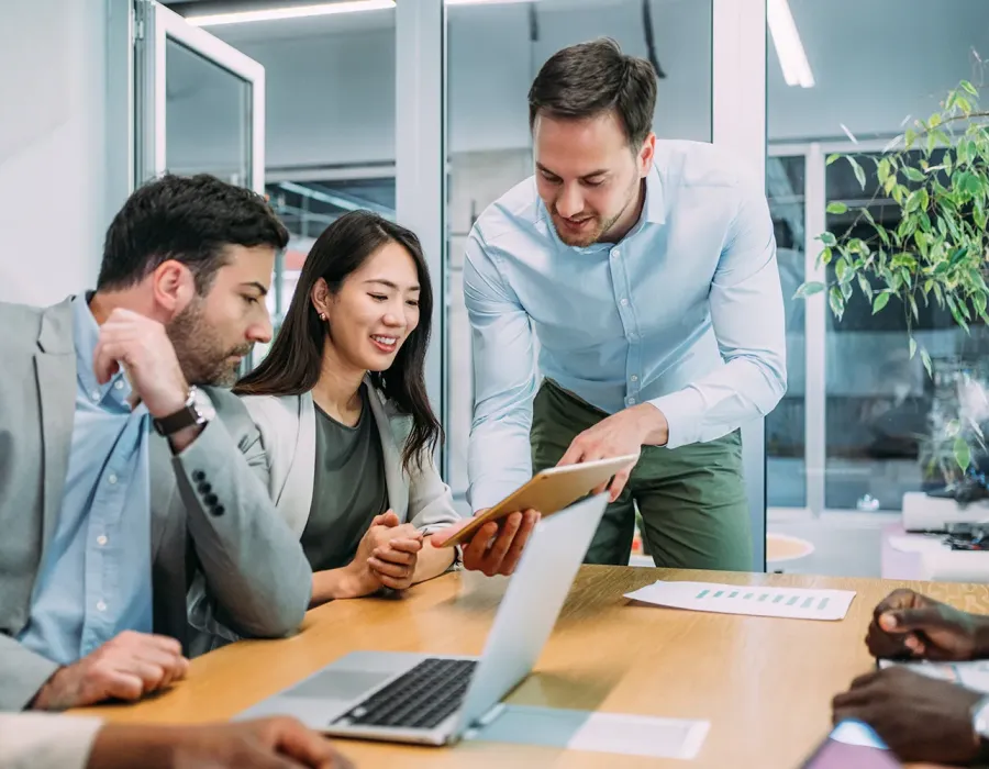 Team of business professionals collaborating at a conference table, reviewing information on a tablet with a laptop and documents in a modern office.